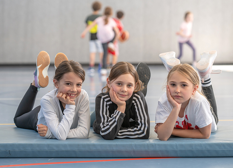 Drei Mädchen beim Schulsport auf einer Matte, im Hintergrund basketballspielende Kinder